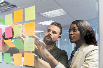 Caucasian man and a african american woman sticking post it on the glass wall in the office