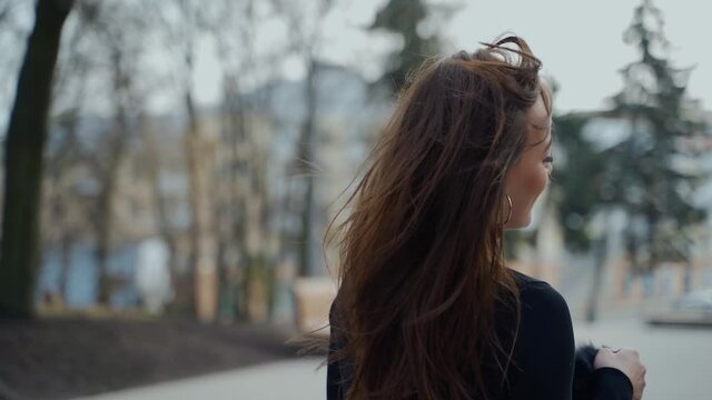 Happy girl turning around in the park. Attractive young lady with long hair in black sweater looking on camera and smiling in the street of a city. Slow motion.