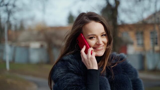 Portrait of a smiling girl with a phone. Beautiful young woman dressed in fur coat seductively poses for camera and talks the phone in the city park. Motion camera around.