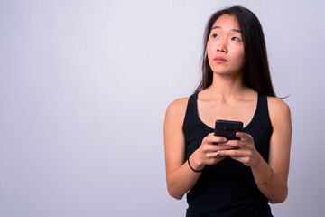 Portrait of young beautiful Asian woman thinking and using phone