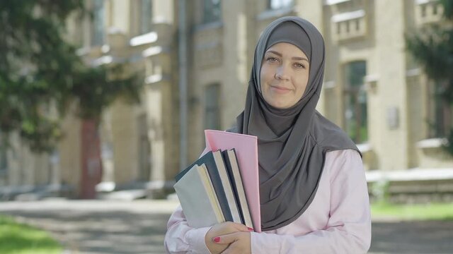 Portrait of happy Muslim woman in grey hijab posing in front of university on sunny day. Positive female immigrant studying in European college. Cheerful young lady with books smiling at camera.