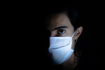 Profile portrait of a man with a disposable white face mask looking aside at the camera with a serious expression. Low key light photography with black background.