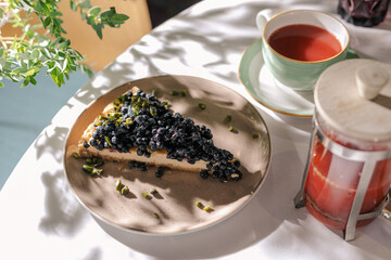 A piece of blueberry pie. Lies on a plate. fruit red tea is poured nearby. The sun is shining on the cake. On a piece, a lot of fresh blueberries. Tea drinking. Dessert in a cafe. Fresh pastries.