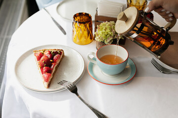 Piece of pie with fresh strawberries. Hot berry tea is poured into a cup. White tablecloth on the table. Tea party. Tea and dessert. Seasonal berries. Dessert in a cafe. Strawberry cake.