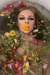 Young woman relaxing during spa in bath full of flowers and holds a yellow flower in his mouth