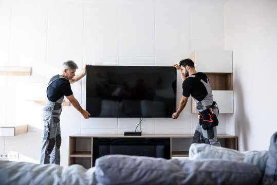 Two professional technicians, workers in uniform installing television on the wall indoors. Construction, maintenance and delivery concept
