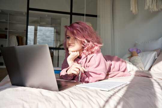 Teenage girl school college student with pink hair watching online course video webinar study in bed. Gen z teen looking at laptop learning lying in bed distance elearning video conference chat call.