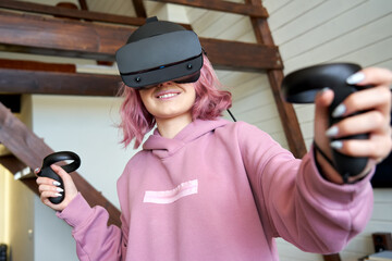 Happy young adult woman gamer teen girl pink hair wear vr headset goggles hold controllers play...