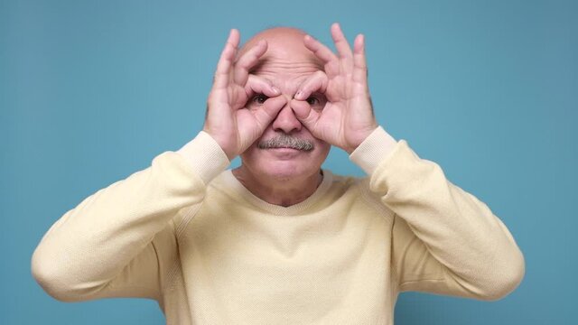 Senior man holding fingers near eyes like glasses or mask like super hero.