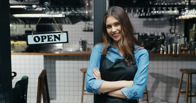 Portrait of beautiful Caucasian young happy woman waitress in apron smiling to camera with table open at cafe entrance outdoor. Pretty cheerful barista standing at bar door with board open.