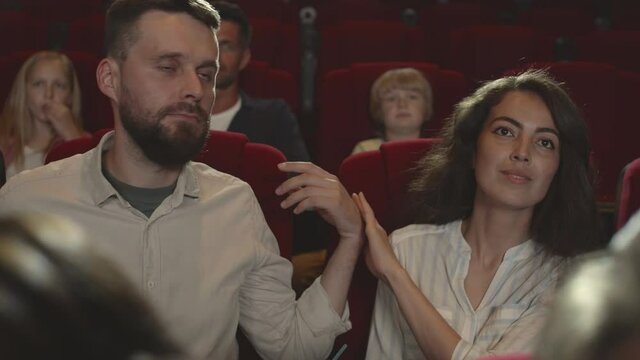 Zoom out of bearded man trying to hug his girlfriend while watching film in cinema but she refusing. The other man embracing his black-haired girlfriend and they enjoying movie