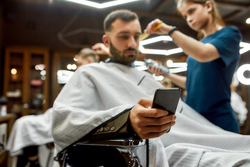 Young handsome bearded man visiting barbershop, sitting in chair and using his smartphone while professional barber girl making trendy haircut for him