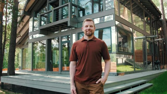 Tracking portrait shot of happy bearded man standing before modern house with panoramic windows and smiling for camera