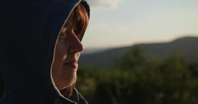 A young woman in a hood looks ahead. In the background of the mountain