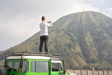 Man visiting Bromo volcano by jeep and standing on the top of the car using a mobile phone