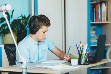 Cute young boy in blue shirt sitting behind desk in his room next to laptop and study. Teenager in earphones makes homework, listening lesson. Home, distance education, self education by  kids.