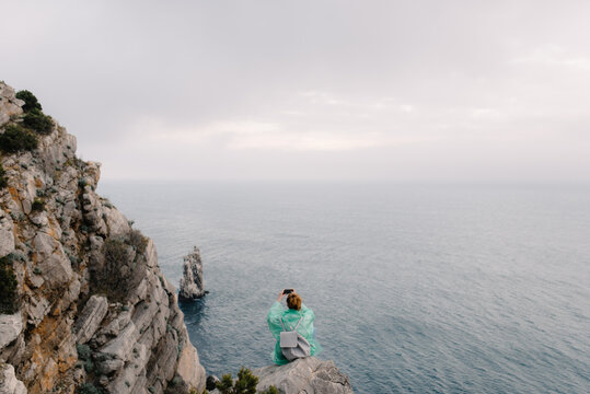 Female sitting on the edge of cliff among the ocean and taking pictures on her smartphone