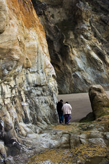young man helping elderly woman walk along slippery and rough ground on rocky beach with high sea cliff walls