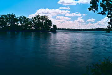 A long exposure of an island full of trees in a river with a smooth waterfront with a background of blue sky with clouds in a summer sunny day