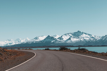 Snowy mountains and large lake after a curve to the left in an asphalt road on a sunny day with blue sky in Patagonia, Chile.