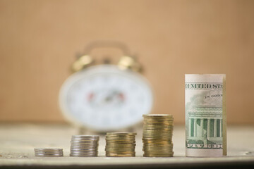 Several stacks of coins with dollar bill and clock in the background out of focus
