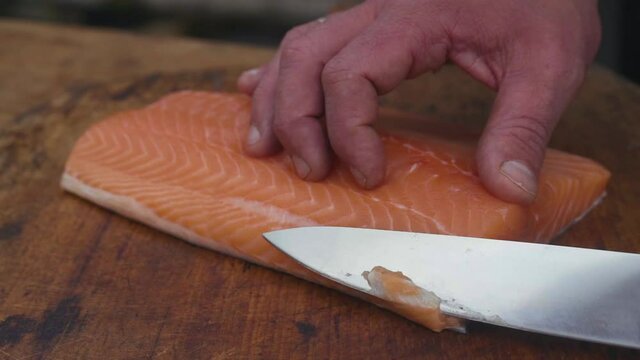 Professional preparation of fish – closeup of man's hands cutting raw piece of salmon with a sharp knife on a wooden plate. SLOW MOTION 50 fps.