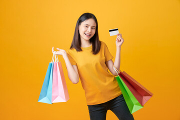 Fototapeta na wymiar Cheerful beautiful Asian woman holding multi coloured shopping bags and credit card on light yellow background.
