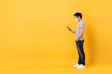 Young Asian man wearing medical face mask standing and using mobile phone isolated on yellow studio background with copy space