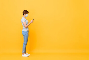 Fototapeta na wymiar Young Asian woman wearing medical face mask standing and using mobile phone isolated on yellow studio background with copy space