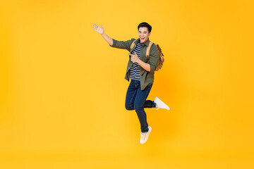 Fototapeta na wymiar Excited smiling Asian tourist man with backpack jumping and pointing hands to empty space aside on isolated yellow studio background