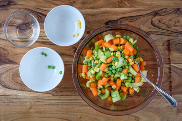 Making a cold salad, glass bowl with chopped fresh and frozen vegetables, and empty bowls, on wood cutting board
