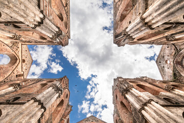 The cross shaped intersection of the transet of an ancient and abandoned church with no roof, as seen from below, under a blue summer sky with puffy clouds