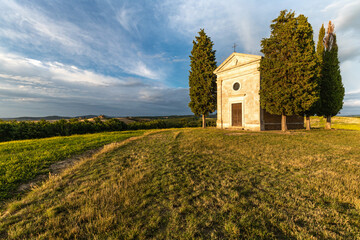 Wide angle view of a hill with rural chapel surrounded by cypresses at sunset, under a blue summer sky with trailing clouds
