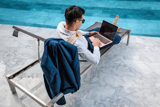 Freelance Concept. A Man With A Laptop In A Warm Climate Works Sitting Near The Pool. Back View.