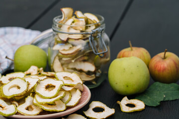 Naklejka na ściany i meble Dried apple slices in open glass jar. Homemade organic dried apple chips with fresh apple on black table background. Sweet vegan snack. Healthy and nutrition concept. Shallow depth of field