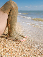 View of beautiful female feet with bright red pedicure on the sand of the beach. The ankles of the feet are smeared with healing mud. Skin rejuvenation while relaxing at sea. Spa. Copy space.