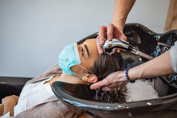 Hairdresser and customer in a salon with medical masks during virus pandemic. Working with safety mask. Hair dresser washing the hair of a client during coronavirus quarantine