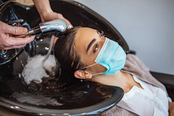 Fototapeta premium A young woman in a medical mask washes her hair in a barbershop during the covid-19 coronavirus pandemic. Reopening with security measures of Hairdressers in the Covid-19 pandemic