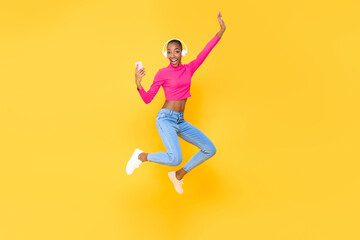 Smiling energetic African American woman wearing headphones listining to music from smartphone and jumping isolated on colorful yellow background