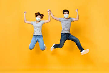 Young Asian couple wearing medical face masks and jumping with arms raised isolated on yellow background, Coronavirus protection concept