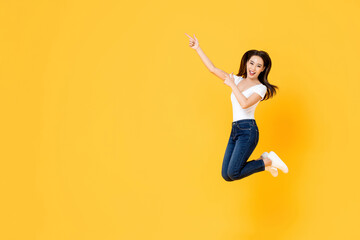 Fototapeta na wymiar Happy smiling young Asian girl jumping with hands pointing up isolated on yellow studio background with copy space