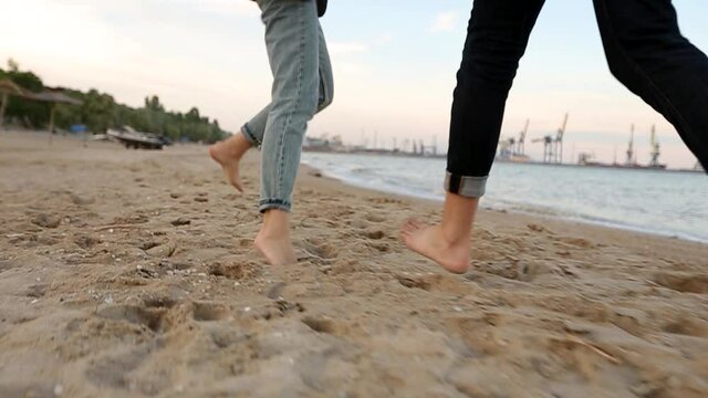 Back low angle view of romantic couple feet running on sand holding hands on beach near sea in slow motion. Boyfriend and girlfriend having fun on ocean shore. Man and woman in love together.
