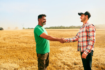 Naklejka na ściany i meble Farmer and agronomist shaking hands in wheat field after agreement. Agriculture business contract concept. Corporate farmer and landlord rancher negotiate with handshake. Combine harvesters harvest.
