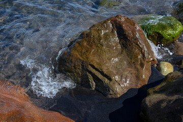 Sea water tide on mossy wet stones and black sand beach. Relaxing natural scene. Marine coast tide foam. Wet rocks on beach closeup. Seaside stone in weed. Coastal day concept. Tropical island holiday