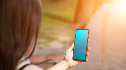 A brunette girl is sitting in the park, holding a mobile phone in her hands. Sunlight, blurred background. lifestyle.