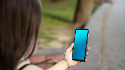 A brunette girl is sitting in the park, holding a mobile phone in her hands. Sunlight, blurred background. lifestyle.