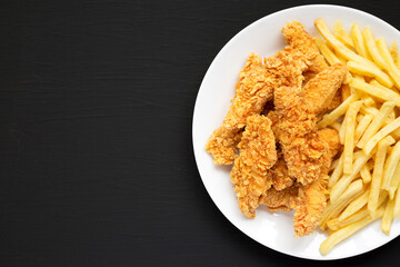 Homemade Crispy Chicken Tenders and French Fries on a white plate on a black background, top view. Copy space.