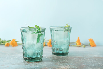 Two beautiful vintage turquoise glasses with cold drink and ice cubes, with fresh green mint leaves decorated with marigold flowers on natural colorful stone table. Summer concept.