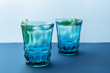 Two beautiful vintage blue glasses with cold drink and ice cubes, with fresh green mint leaves on deep blue and light blue background. Backdrop for your design.
