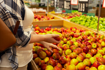 Young woman in a supermarket. Woman holding red apples while choosing fresh fruits and vegetables at farmers market, copy space